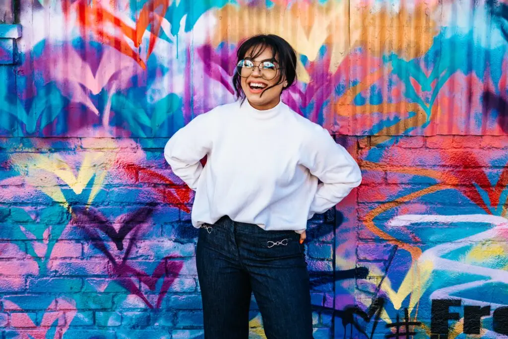 Smiling young woman in white sweater and glasses standing in front of a colorful graffiti wall.