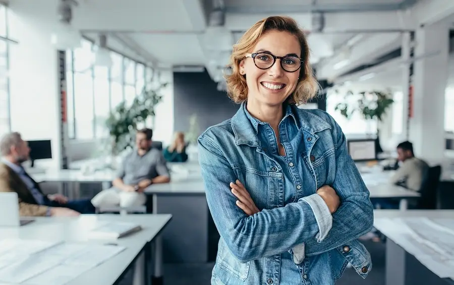 Smiling woman in denim jacket with arms crossed standing in a modern office with colleagues working in the background.