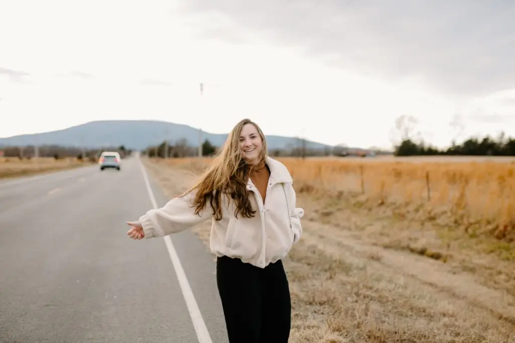 Smiling young woman with long hair standing on a rural roadside with mountains and open fields in the background.
