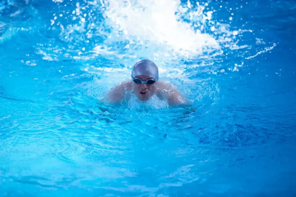 Swimmer in cap and goggles surfaces through splashing blue pool water during a stroke.
